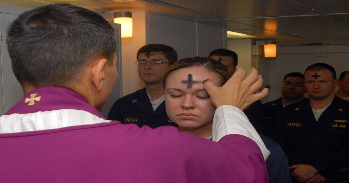 Picture of Ash Wednesday service. Foreground: back view of clergy in maroon robe with cross on collar, applying an ash cross on the forehead of a woman. Background: faces of various men with ash crosses on forehead and wearing Army uniforms.